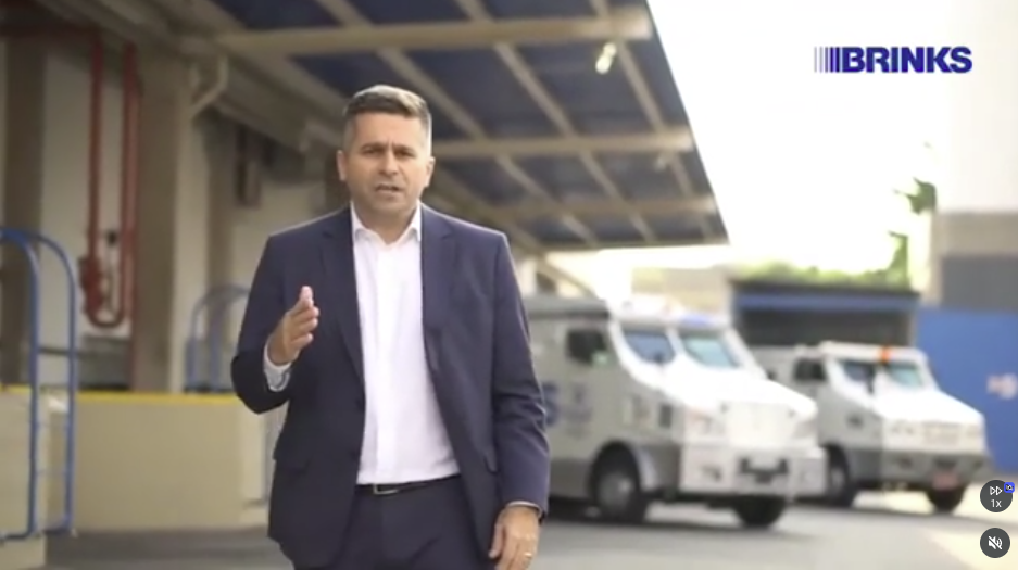 Apresentador da Brinks em pátio com carros-fortes ao fundo